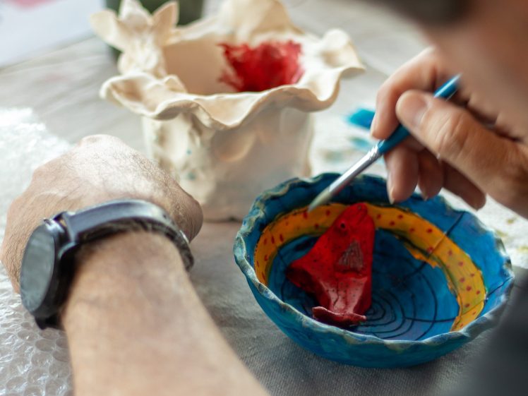 Image of A person wearing a watch paints a red ceramic object inside a colourful bowl on a table, with another unpainted ceramic piece and painting materials nearby.
