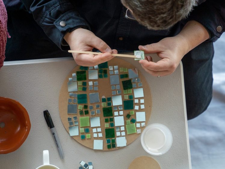 Image of A person arranges small coloured tiles on a round base, creating a mosaic pattern. Their hands hold a stick and a tile. Various tiles, glue, a marker, and a bowl are on the table beside them.