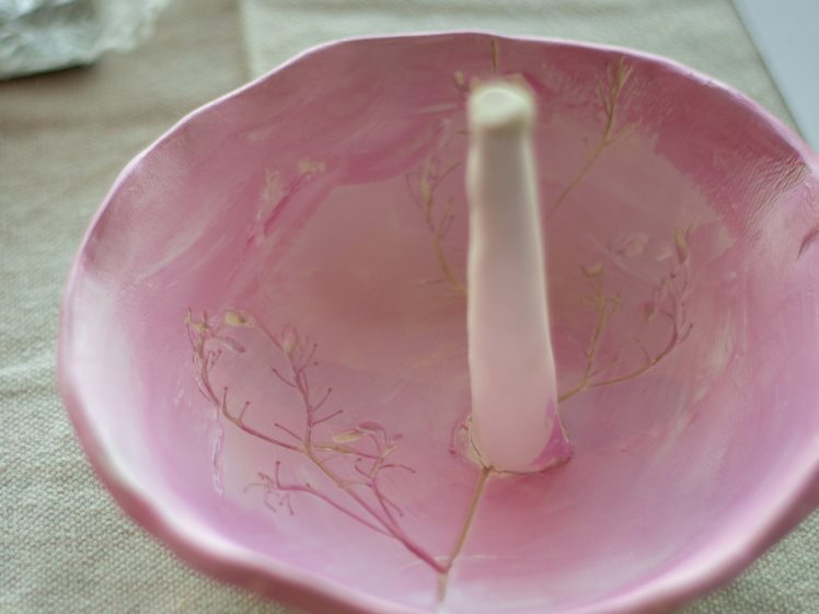 Image of A pink ceramic dish with a central handle, featuring delicate leaf imprints, sits on a beige fabric surface under soft light.