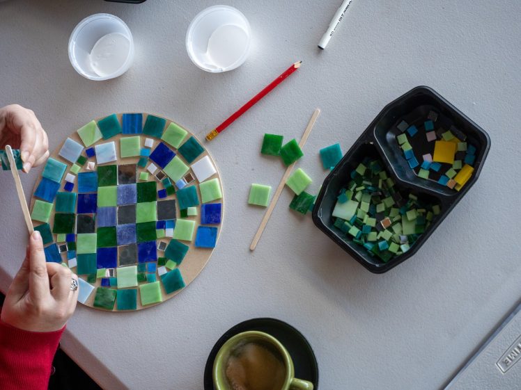 Image of A person arranges square glass tiles on a circular mosaic, with various green and blue tiles, two cups, a pencil, a coffee mug, and a marker on a grey table.