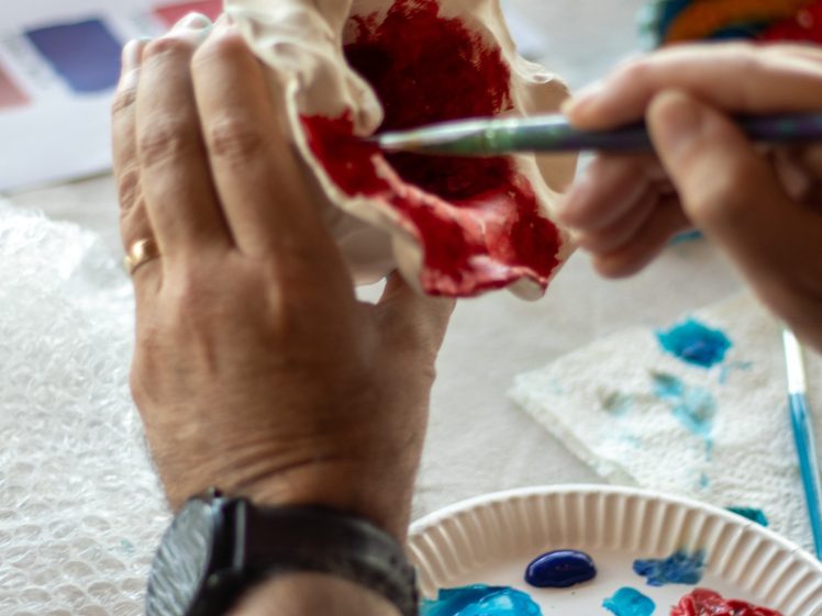 Image of A person paints the inside of a white ceramic object red, using a brush. Their palette holds various blue, yellow, and red paints. A colour sample sheet and bubble wrap are also visible on the table.
