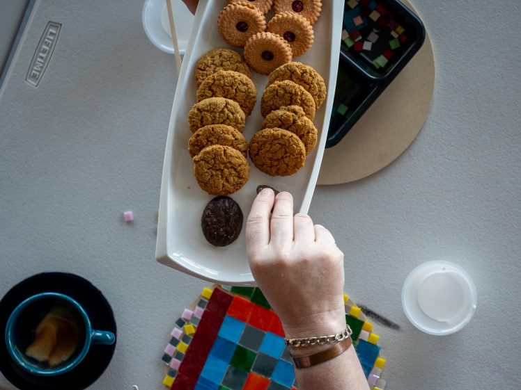 Image of A hand reaches for a rectangular plate of assorted biscuits above a table set with a cup of coffee, colourful mosaic tiles, and small containers.