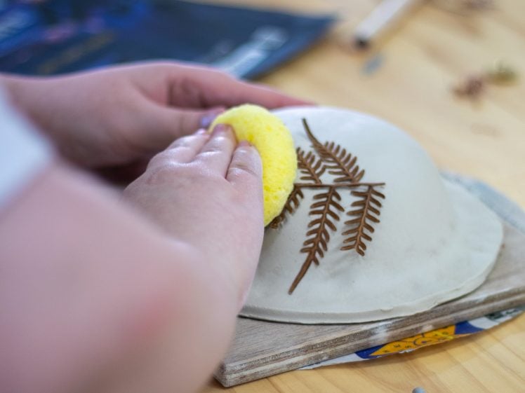 Image of A person uses a yellow sponge to smooth the surface of a clay bowl decorated with impressed fern leaf patterns on a wooden table. Art tools and materials are visible in the background.