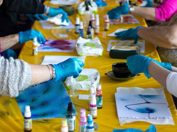 Image of People wearing blue gloves work on colourful art projects at a long table covered with a yellow tablecloth, using paint, brushes, and paper. Bottles of ink and art supplies are scattered across the table.