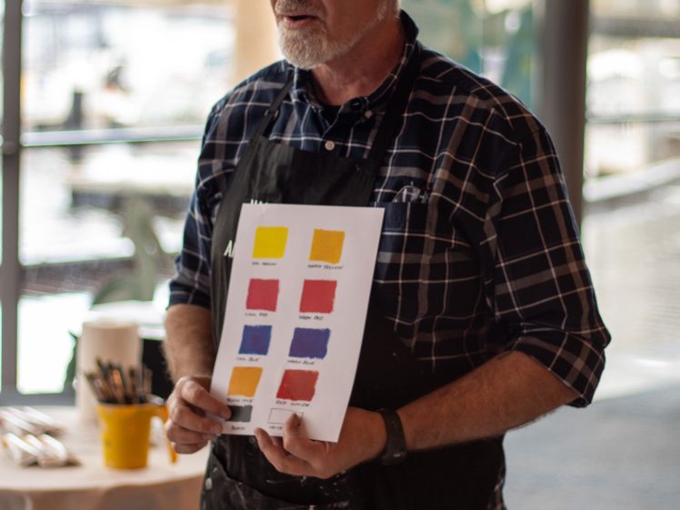 Image of A man in a checked shirt and black apron holds up a sheet with colourful paint swatches in a bright room, possibly during an art class or workshop. Art supplies and a yellow cup are visible on the table beside him.
