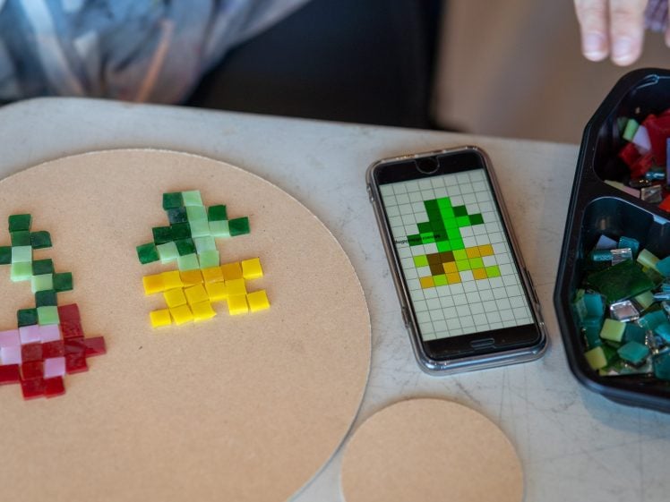 Image of A round board with two pixel art flower designs made of coloured tiles sits on a table next to a phone displaying a similar design and a container of mosaic tiles. A person's hands are visible nearby.