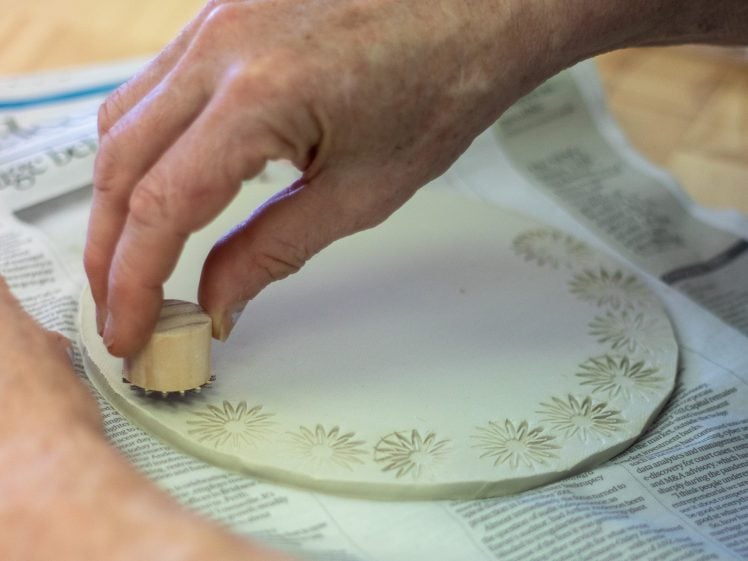 Image of A person presses a small wooden stamp into the edge of a round piece of clay, creating a floral pattern. The clay rests on a sheet of newspaper on a table.
