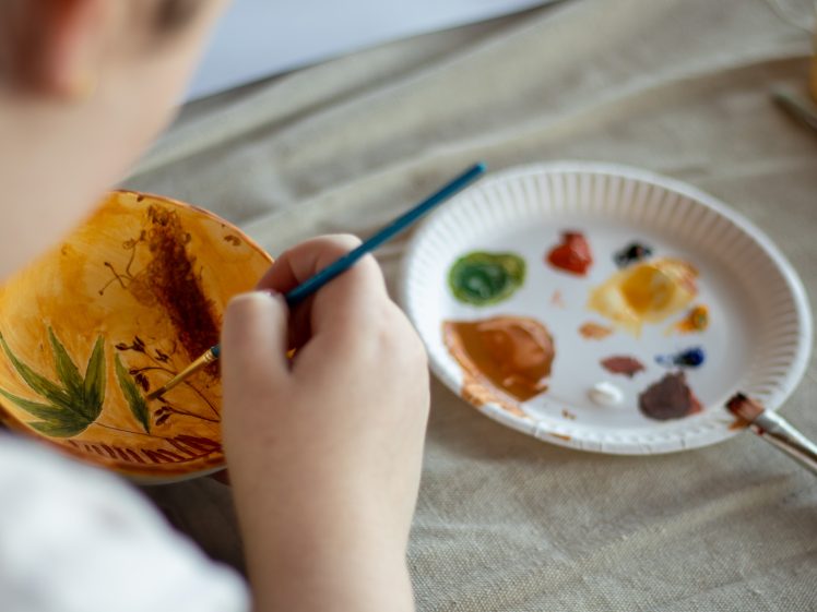 Image of A person paints a ceramic bowl with a blue brush, using various colours from a white paper palette nearby. The bowl features green and brown designs, and the scene is set on a beige cloth surface.