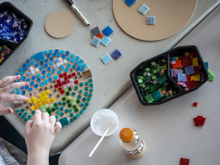 Image of A person arranges colourful glass mosaic tiles on a circular board. Containers hold blue, green, and mixed tiles nearby, along with glue and a stick, on a white table.