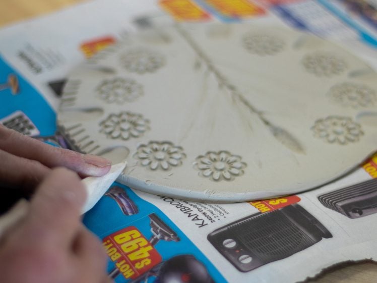 Image of A person shapes the edge of a round clay slab decorated with flower patterns, placed on a colourful newspaper on a table.