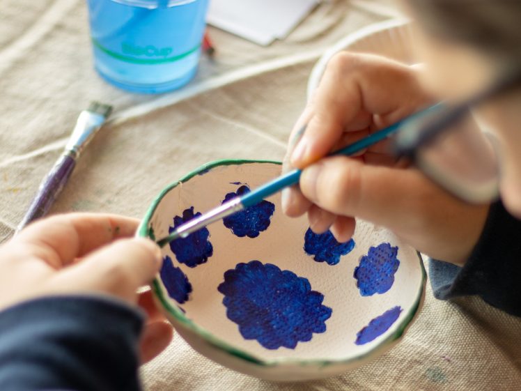 Image of A person paints blue floral patterns on a white ceramic bowl using a fine brush. A cup with blue water, a paintbrush, and paper are visible on the table.