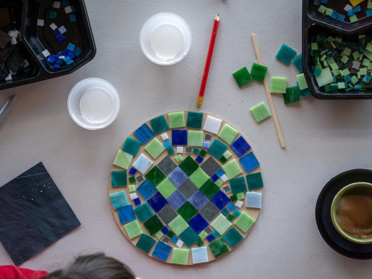 Image of A circular mosaic in progress using green, blue, and cream tiles is on a table surrounded by containers of tiles, glue, a pencil, a lolly stick, a coffee cup, and a black serviette.