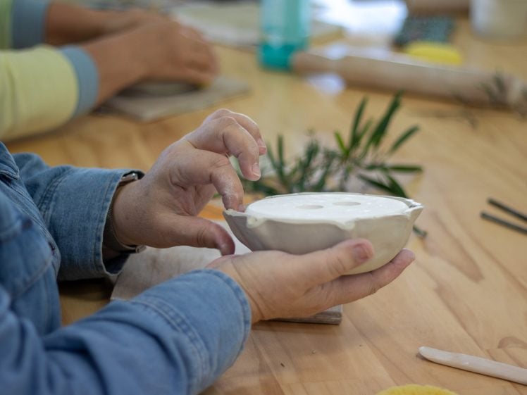 Image of A person shapes a clay bowl with their hands on a wooden table, surrounded by pottery tools and leaves. Another person works in the background.