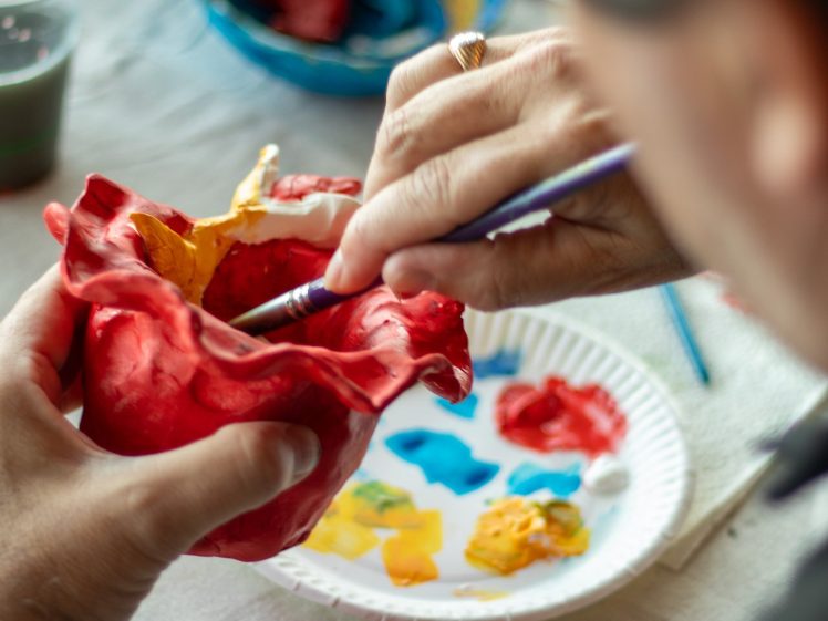 Image of A person paints a red heart-shaped object with yellow paint using a brush. A palette with various paint colours and cups are on the table in the background.