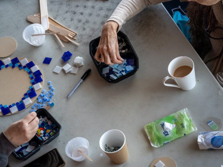 Image of Two people sit at a table assembling a blue tile mosaic. Assorted mosaic tiles, wooden sticks, cups, a marker, a tissue packet, and drinks are scattered on the table. One person reaches into a container of tiles.