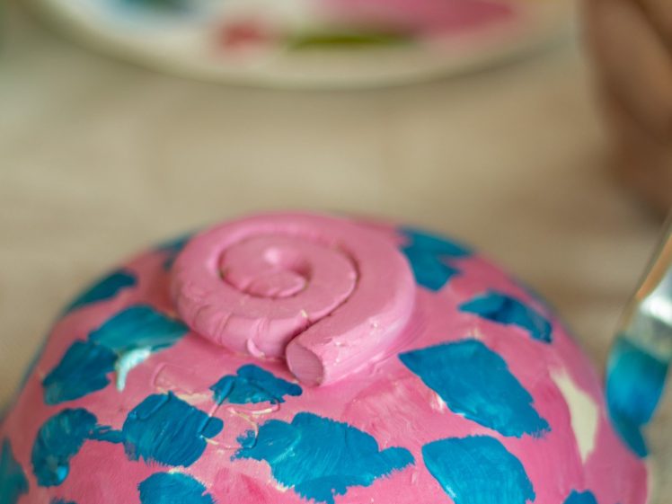 Image of A hand paints blue spots onto a pink, spiral-shaped ceramic bowl. A blurry palette with green and pink paint is visible in the background.