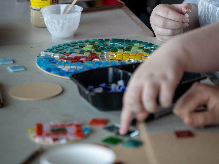 Image of Two people work together on a mosaic art project, arranging small colourful glass tiles on a circular board. Various tiles and materials are scattered on the table.