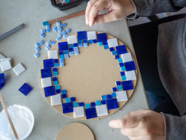 Image of A person is arranging blue, white, and turquoise square mosaic tiles in a circular pattern on a round board, with craft supplies and glue visible on the table.