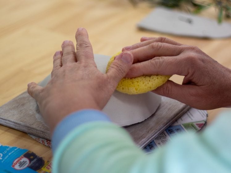 Image of A person uses a yellow sponge to smooth the surface of a white clay object on a wooden board, with various materials and a newspaper visible on the table.
