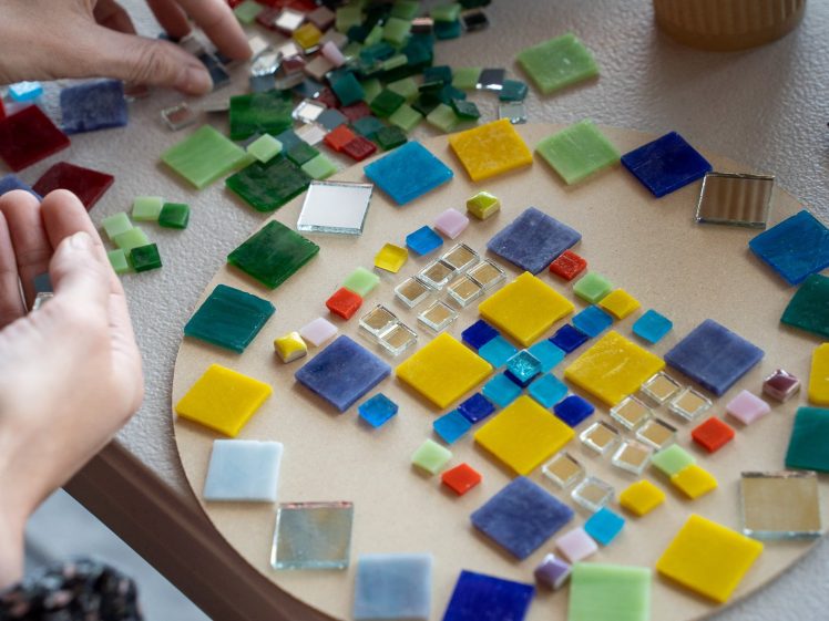 Image of Hands arranging colourful glass tiles into a geometric mosaic pattern on a round board, with various loose tiles scattered on the table nearby.