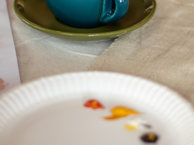 Image of A teal coffee cup with a saucer sits on a table next to a paper plate with blobs of colourful paint, suggesting an artistic workspace.