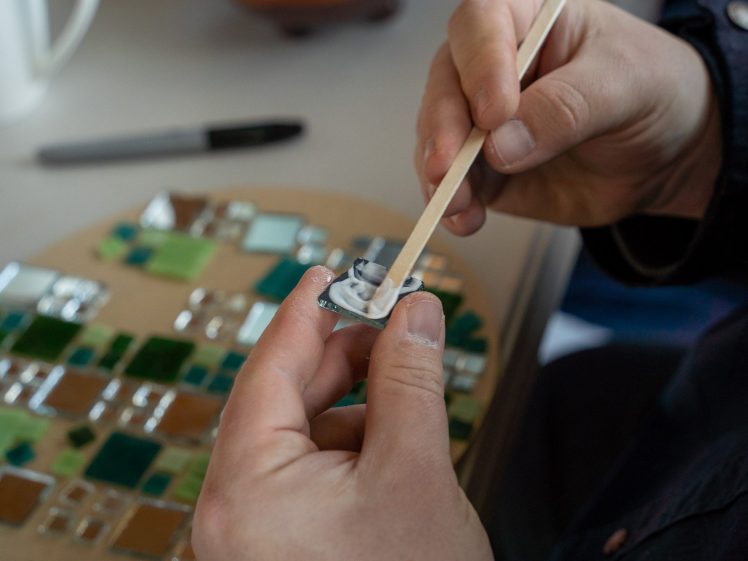 Image of A person uses a wooden stick to apply white adhesive to the back of a small glass tile, with a mosaic of green and brown tiles visible in the background.