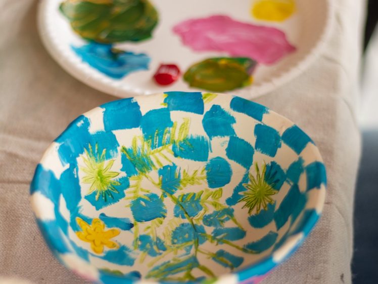 Image of A hand-painted ceramic bowl with blue chequered and yellow-green floral patterns sits in front of a paper plate decorated with colourful paint splodges, all on a beige fabric surface.