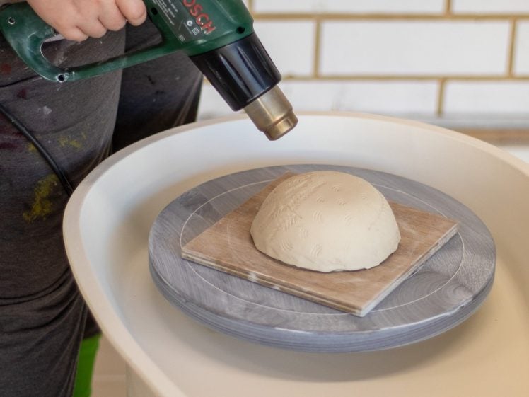 Image of A person uses a green heat gun to dry a dome-shaped piece of clay placed on a rotating potter's wheel. The person is wearing grey trousers and bright green shoes. White tiles are visible in the background.