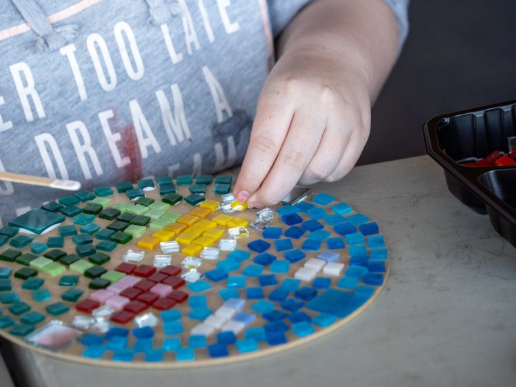 Image of A person arranges colourful mosaic tiles on a round board, creating a vibrant design. Their hand holds a small yellow tile, and a tray with more tiles sits nearby on the table.