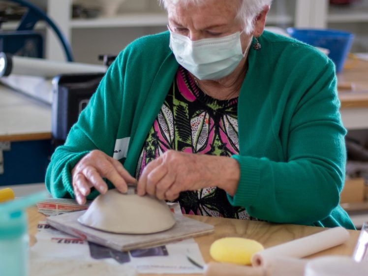 Image of An elderly woman with short white hair, wearing a mask and a green cardigan, shapes clay on a table in a pottery studio. Various art materials and tools are scattered around her workspace.