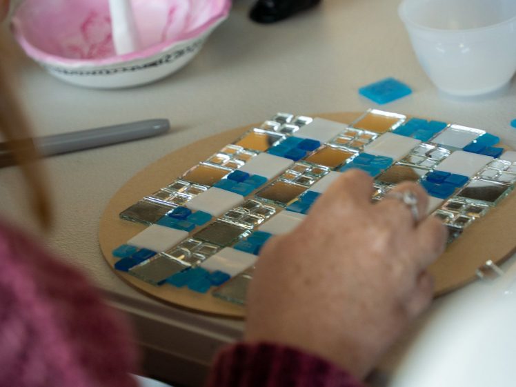 Image of A person creates a mosaic by arranging blue, white, and silver glass tiles on a round board. Nearby are a bowl, spatula, and small cups, indicating an arts and crafts activity.
