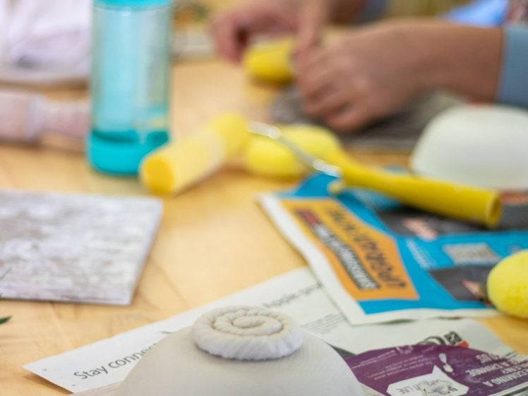 Image of People work on clay projects at a table covered with newspapers, tools, sponges, and a water bottle. A ceramic bowl with a spiral design on top is in the foreground, partially finished.