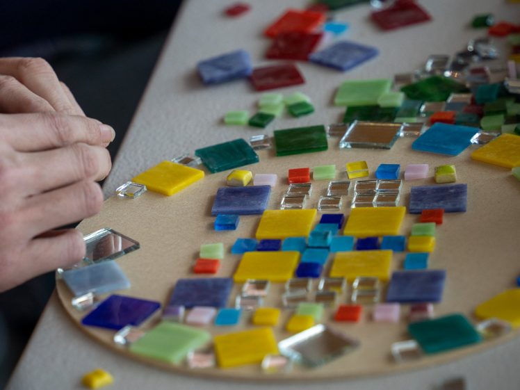 Image of A person arranges colourful glass tiles on a circular board, creating a mosaic design. Various square and rectangular tiles in blue, yellow, red, green, and clear colours are scattered on the table.