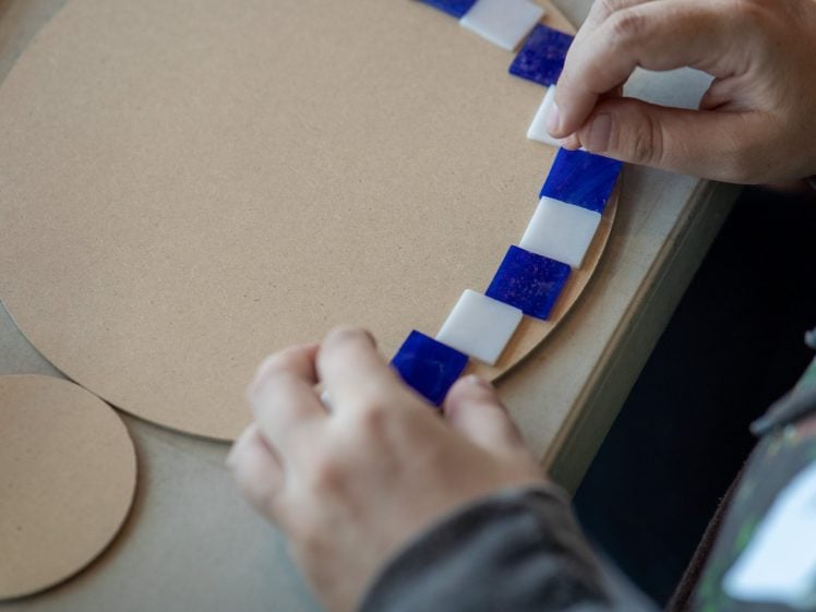 Image of A person arranges alternating blue and white square tiles along the edge of a round, unfinished wooden board, likely preparing a mosaic design.