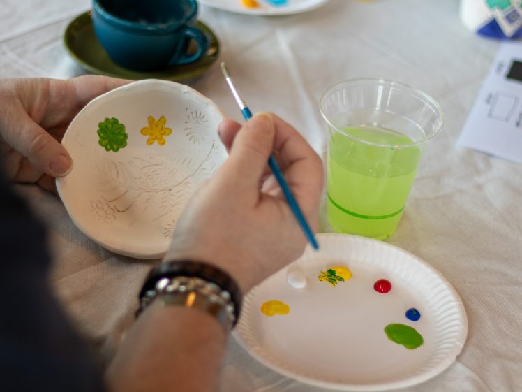 Image of A person paints small flowers on a white ceramic bowl using a fine brush, with paint colours on a paper plate and a cup of green rinse water nearby on a table.