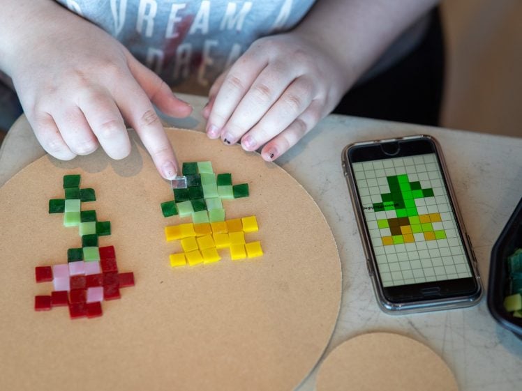 Image of A person arranges coloured mosaic tiles on a round board, following a pixel art flower design displayed on a mobile phone next to them. Their finger points to a green tile on the board.