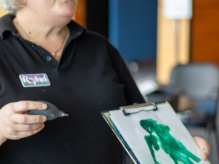 Image of A woman with short grey hair, wearing a black shirt and name tag, holds a paintbrush and a clipboard displaying a green abstract watercolour painting. She appears to be explaining her artwork.