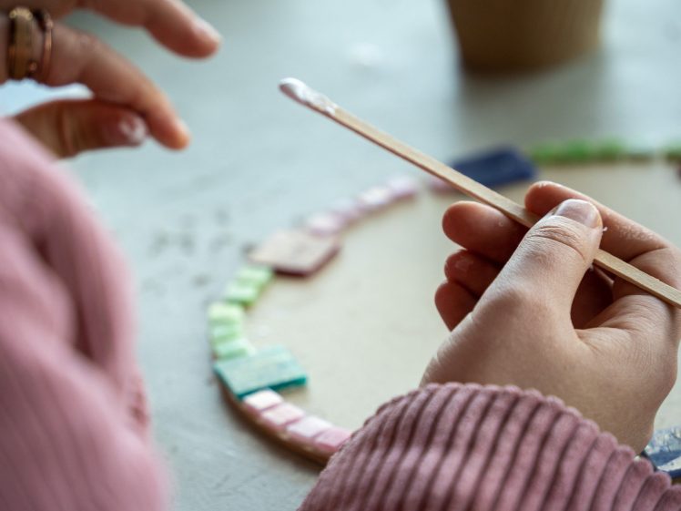Image of Close-up of a person in a pink jumper applying glue to a wooden stick, creating a mosaic with colourful tiles on a round surface.