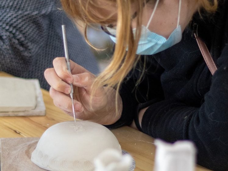 Image of A person wearing glasses and a face mask carefully carves or sculpts a white ceramic bowl, using a pointed tool whilst leaning over a wooden table. Other ceramic pieces and materials are visible nearby.
