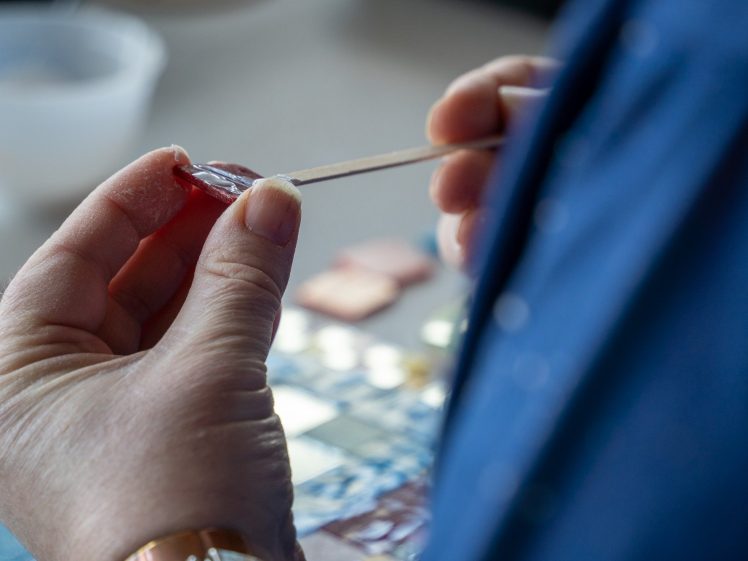 Image of A person holds a small stick and applies a red substance to a piece of material, possibly for a craft or art project, with tiles and a cup visible in the background.