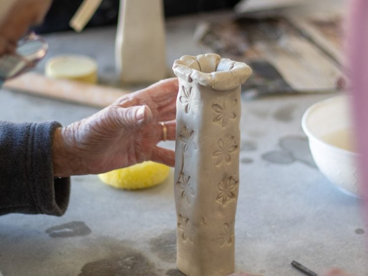 Image of Two people sculpt a tall, flower-patterned vase from clay at a table, with tools and a bowl nearby. One person supports the vase whilst another shapes it, focusing on fine details.