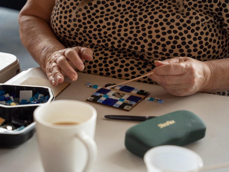 Image of A person wearing a spotty shirt arranges small blue and white mosaic tiles on a board, using a wooden stick. A cup of coffee, pen, glasses case, and a container of tiles are on the table.