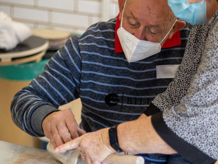 Image of An elderly man wearing a face mask works with clay as a woman, also masked, guides his hands and helps him roll the clay on a table. Both are focused on the activity in a well-lit indoor setting.