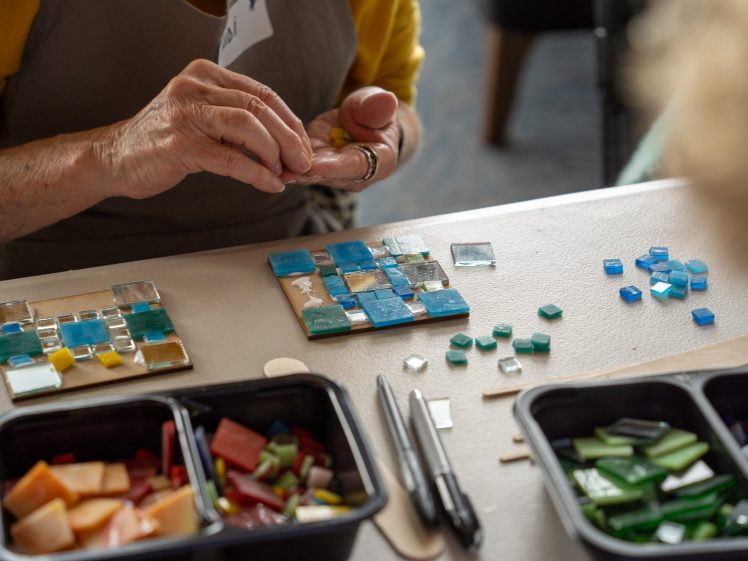 Image of A person arranges blue and green glass tiles on a small board, creating a mosaic. Nearby are trays of colourful tiles and markers on a table. Only the person's hands and arms are visible.