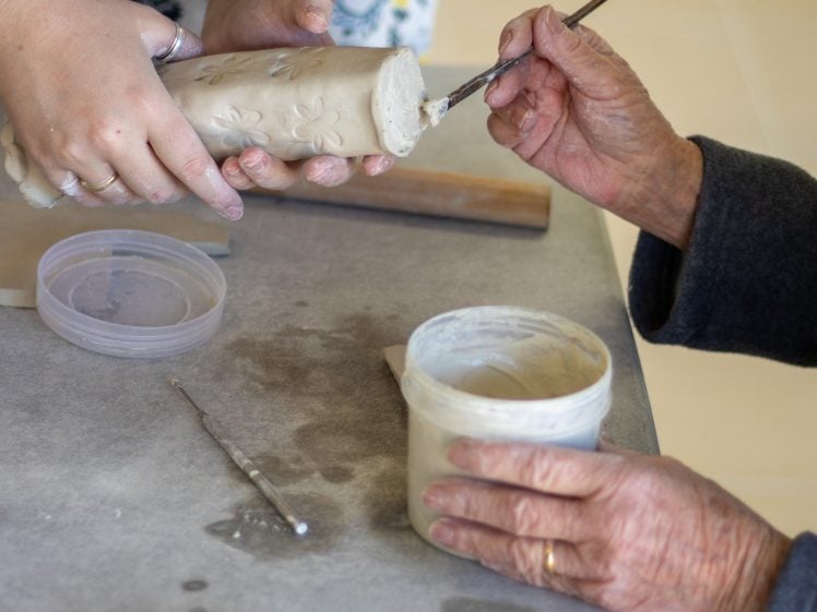 Image of Two people work together on a pottery project, one holding a clay piece with floral designs whilst the other applies slip with a brush. Tools and a sponge are on the table.