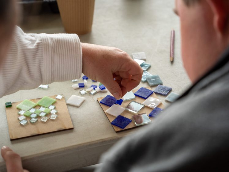 Image of Two people work on a mosaic art project, placing small coloured glass tiles onto wooden boards. One hand is arranging blue and white tiles, while the other board has green and white tiles. A pencil and cup are in the background.
