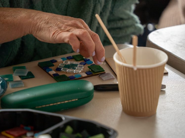 Image of A person’s hand arranges colourful mosaic tiles on a small board. Nearby are a ribbed paper cup with two wooden sticks, a green glasses case, and assorted tiles on a cream-coloured table.