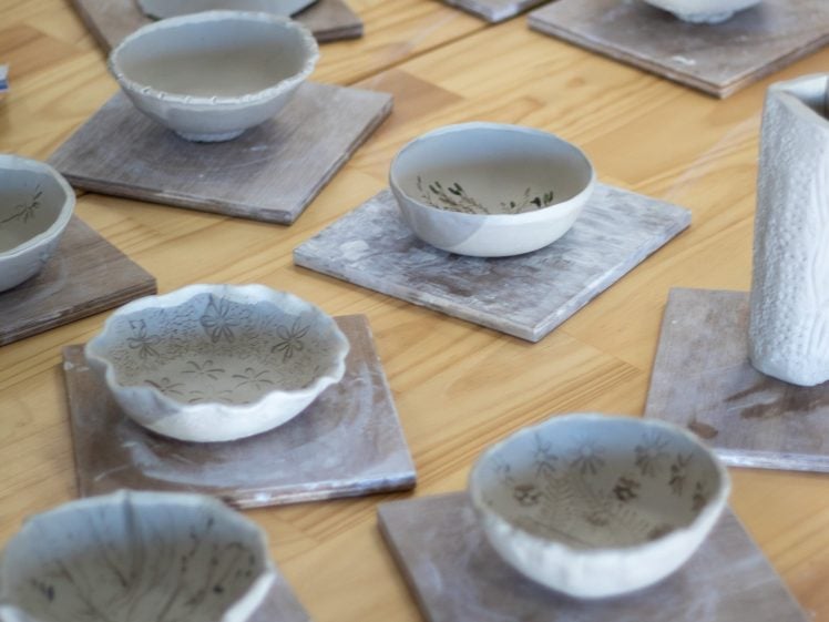 Image of Handmade ceramic bowls and dishes with delicate patterns are displayed on square wooden boards atop a light wood table. Some pieces have textured edges or decorations, highlighting unique craftmanship.