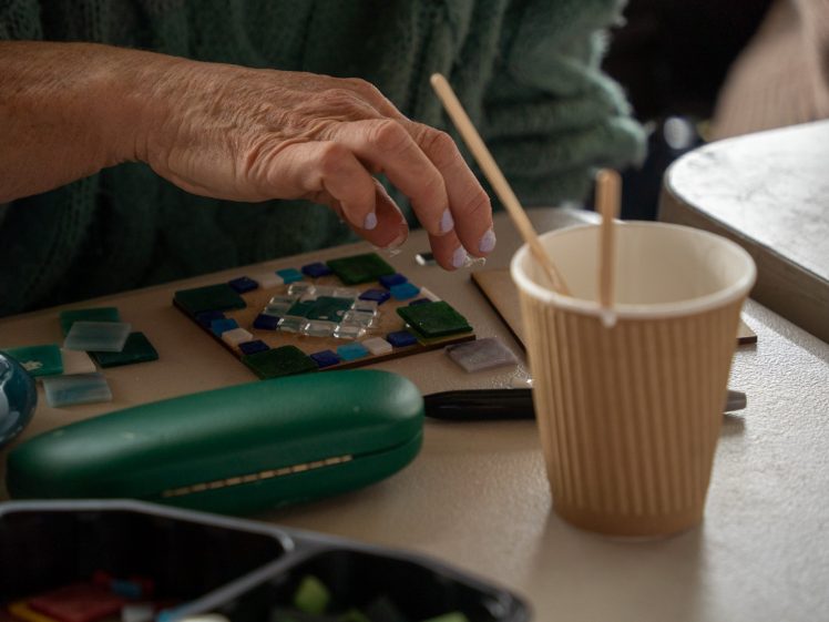 Image of A person’s hand arranges small coloured tiles on a mosaic project. Nearby are a green glasses case, a corrugated paper cup with wooden sticks, and other art supplies on a table.