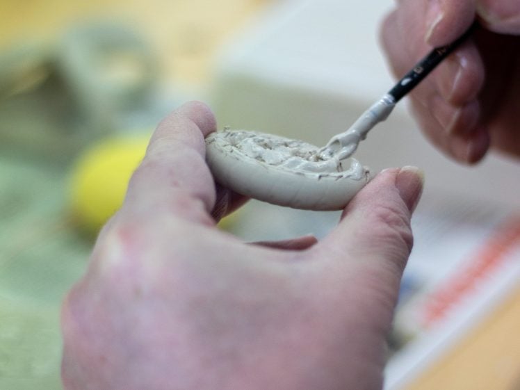 Image of A person uses a small paintbrush to add details to a round piece of clay, holding it carefully in their other hand. The background shows blurred objects on a table.